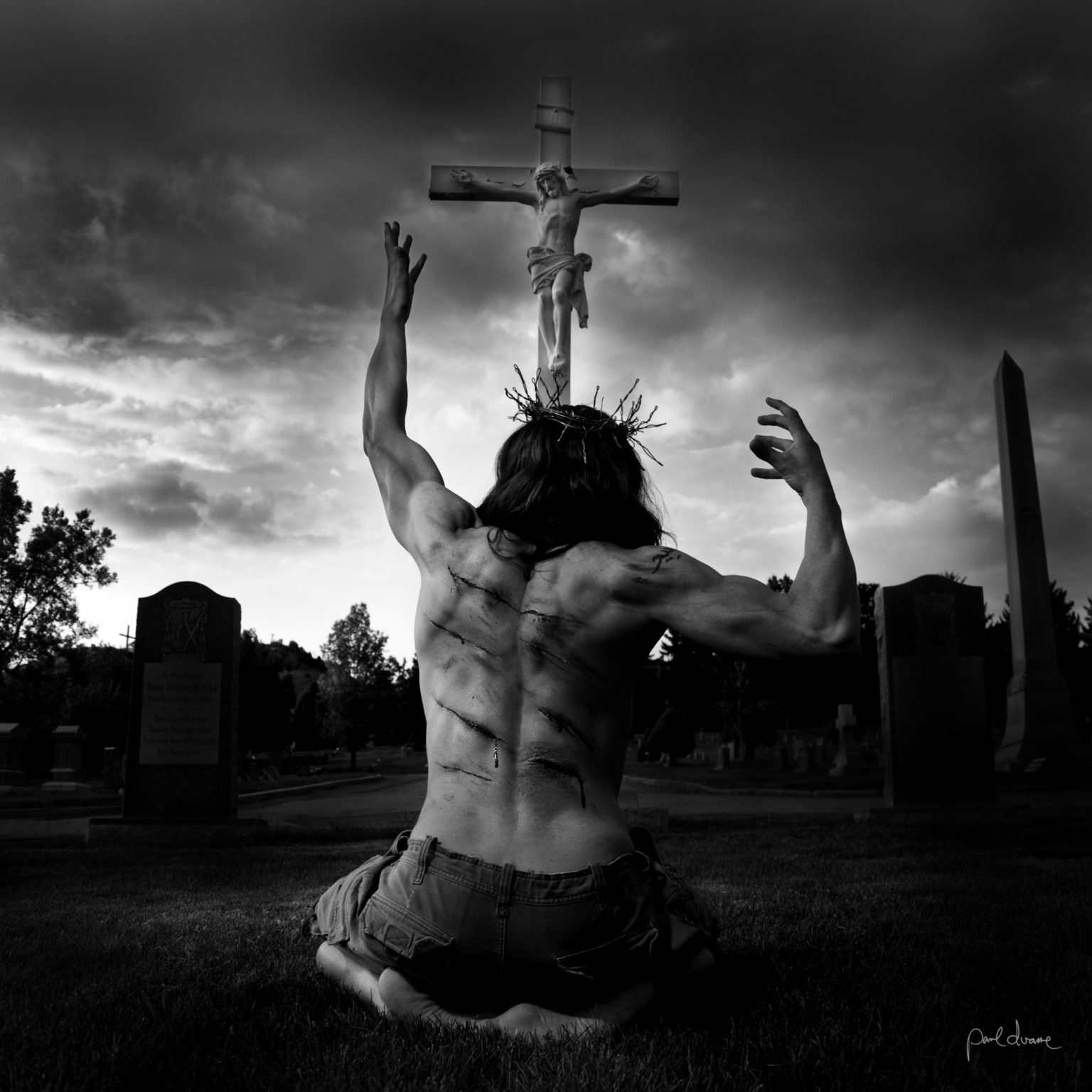A fine art black & white photograph by Paul Duane, depicting a modern Jesus kneeling in suffering before a crucifix.