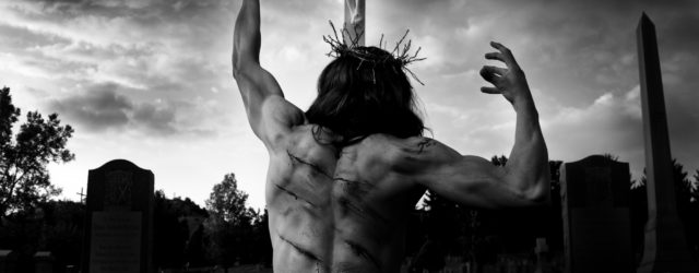 A fine art black & white photograph by Paul Duane, depicting a modern Jesus kneeling in suffering before a crucifix.